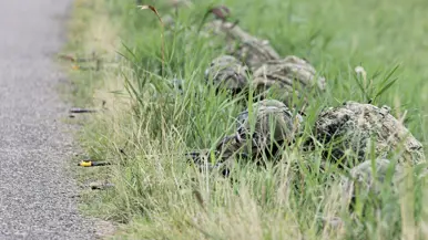 Militairen met helm en uitrusting liggen in hoog gras langs een asfaltweg, omringd door groen en onkruid.