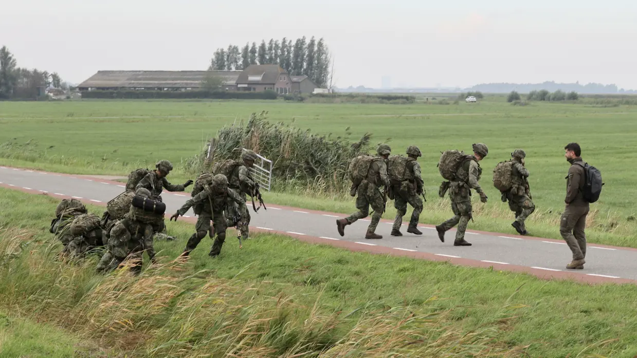 Soldaten in camouflage lopen en kruipen over een landweg door groen landschap met boerderijen op de achtergrond.