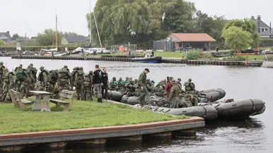 Groep soldaten in camouflage bij boten aan een waterkant, met bomen, huizen en een jachthaven op de achtergrond.