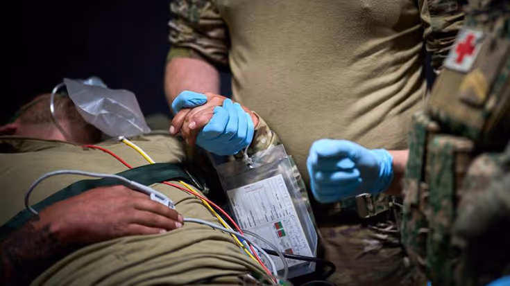 Een militair met blauwe handschoenen houdt de hand vast van een liggende collega die gewond is.