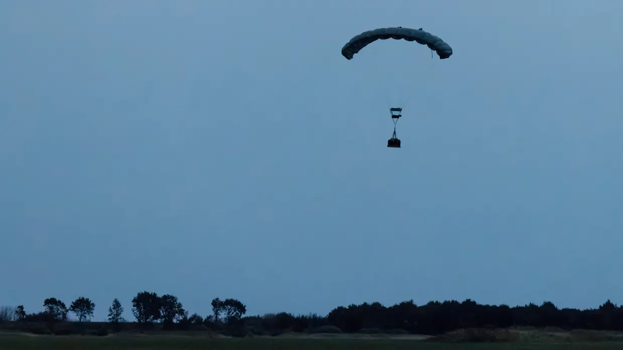 Een lading hangt onder een parachute boven een open veld. 