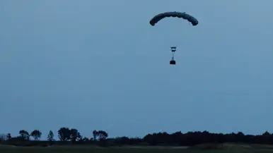 Een lading hangt onder een parachute boven een open veld. 