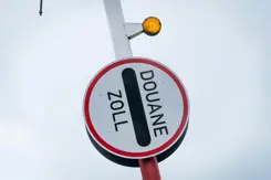 Rechtopstaande slagboom met rond bordje eraan vast, waar de tekst Douane Zoll op staat.
