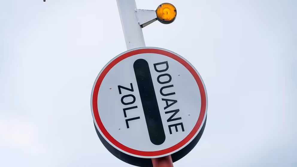 Rechtopstaande slagboom met rond bordje eraan vast, waar de tekst Douane Zoll op staat.