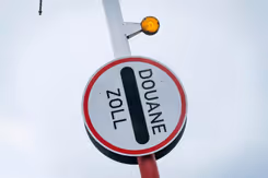 Rechtopstaande slagboom met rond bordje eraan vast, waar de tekst Douane Zoll op staat.