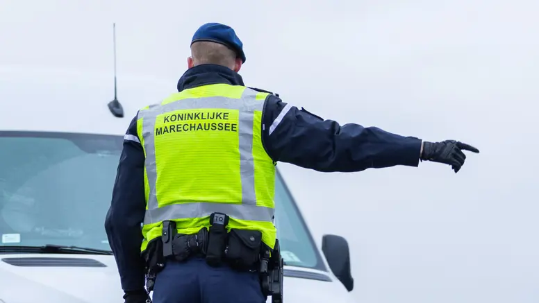 Foto van een marechaussee met een geel hesje. Hij wijst een auto de weg.