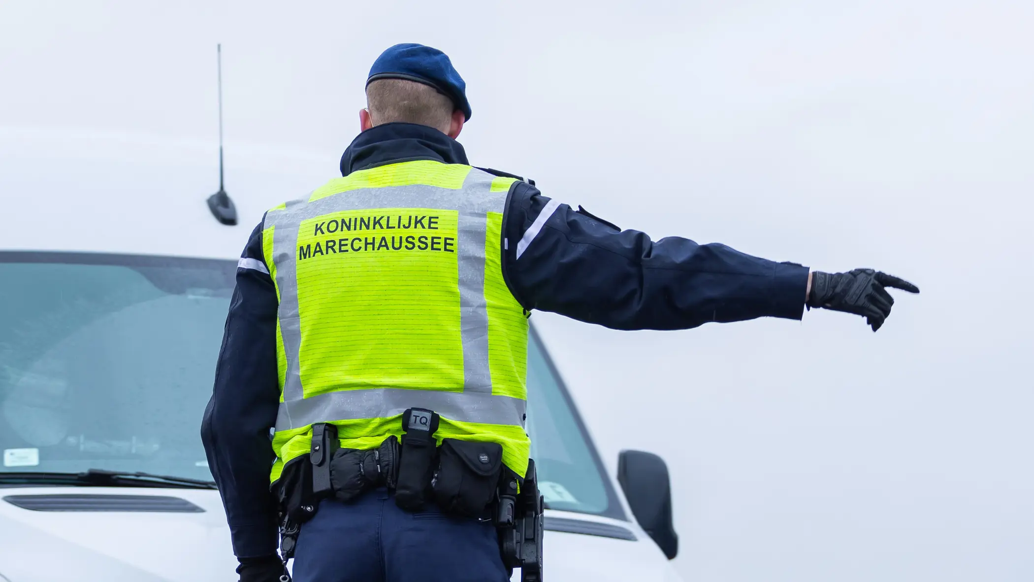 Foto van een marechaussee met een geel hesje. Hij wijst een auto de weg.