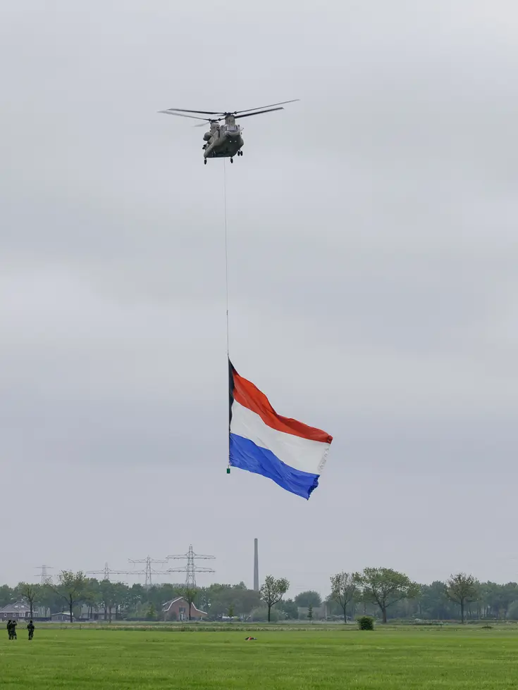 Een Chinook stijgt op boven een veld, met aan een lange kabel de Nederlandse vlag