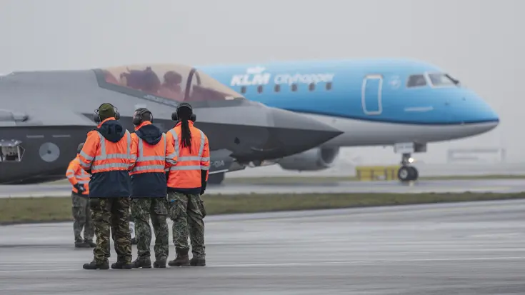 Militair personeel in oranje jassen bij een F-35 op een platform, met een KLM-vliegtuig op de achtergrond.