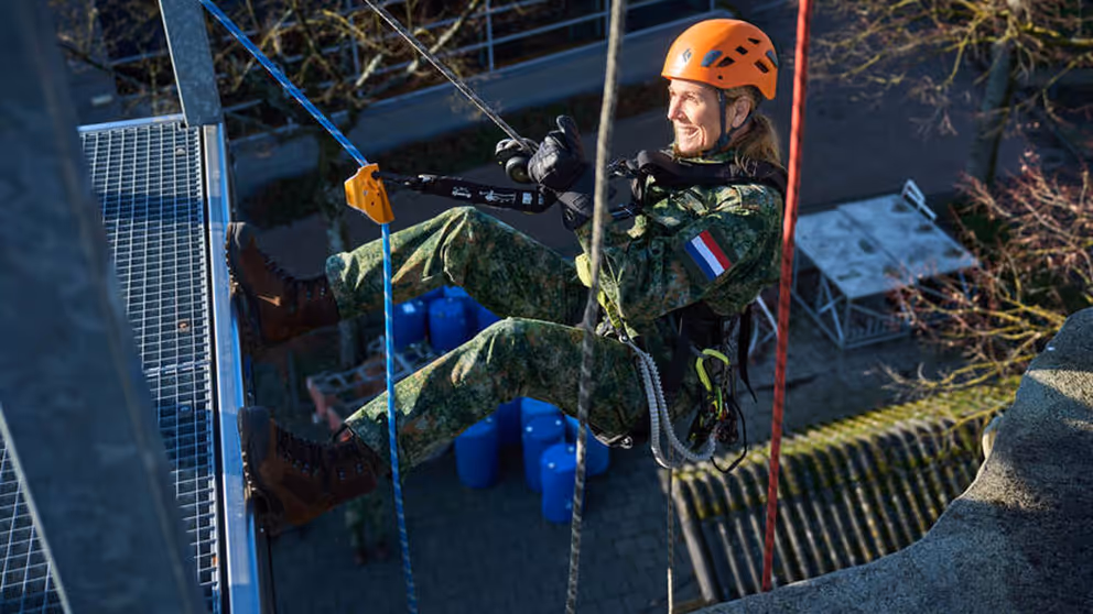Koningin Maxima hangt aan touwen tijdens een oefening en heeft een oranje helm op.