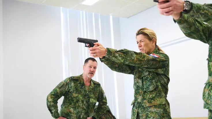 Koningin Maxima heeft een wapen in haar hand en richt het ergens op. Naast haar nog een arm die een wapen vasthoudt.