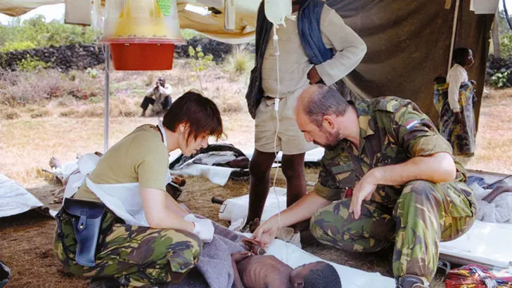 2 militairen van een medisch team verzorgen een vluchteling die met een infuus op een brancard ligt in een vluchtelingenkamp.
