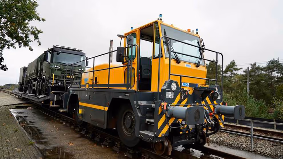 Een gele locomotief trekt groene Defensievrachtwagens.