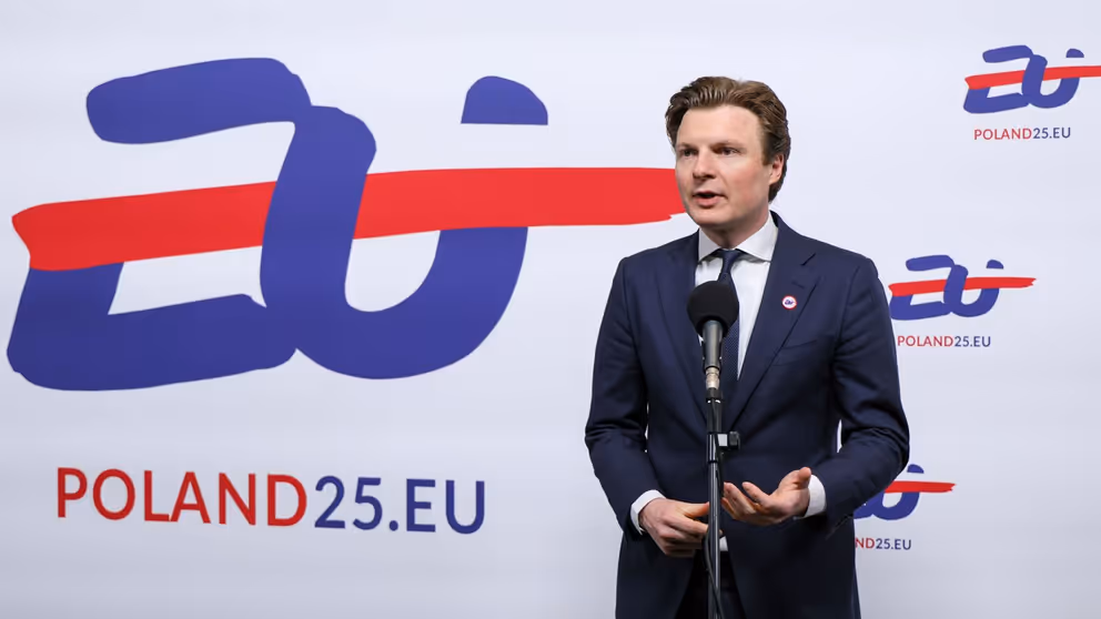 Minister Brekelmans voor een wit bord met blauwe en rode tekst: EU Poland25.EU