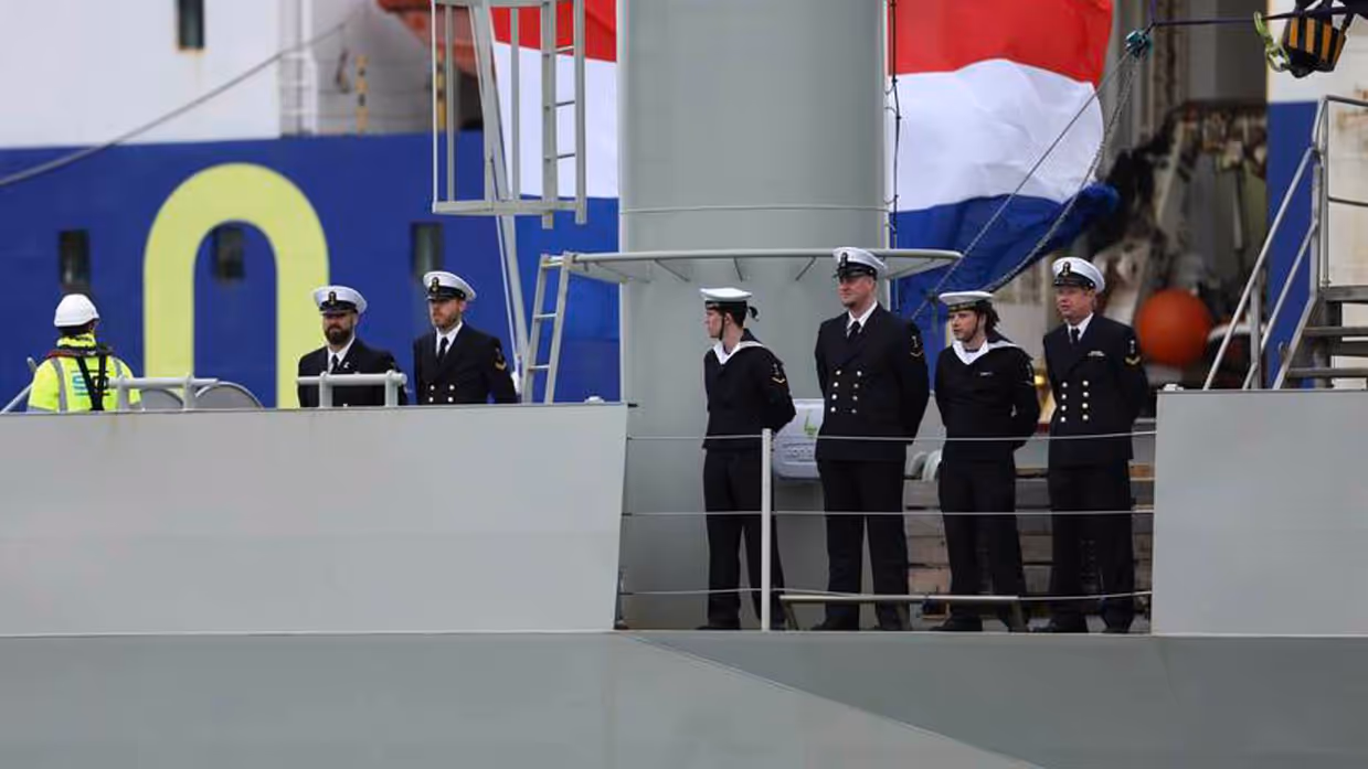 Bemanning staat op het dek. Op de achtergrond de Nederlandse driekleur.
