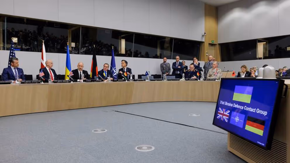 Zaal met sprekers van de 31e Ukrain Defence Contact Group. Op de voorgrond is veel grijze vloer zichtbaar.