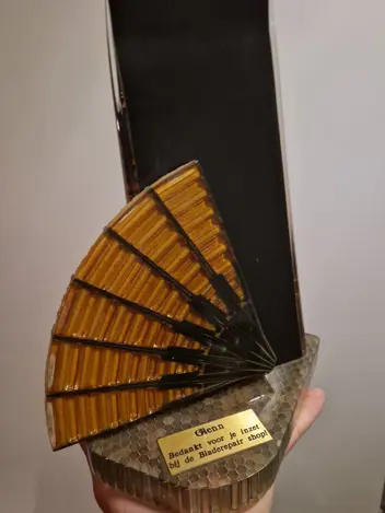 Kunstwerk bestaande uit een waaier tegen een zwart blad, geplaatst op een bruin vlak met honingraatstructuur. Erop een plaatje met de tekst ‘Glenn, bedankt voor je inzet bij de Bladerepair shop!’. Het werk wordt getoond op een handpalm.
