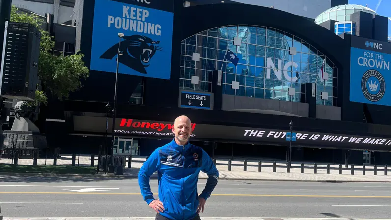 Een glimlachende, kalende man in blauwe sportjas staat trots voor het Bank of America Stadium met ‘Keep Pounding’-borden en lege straten op de voorgrond.