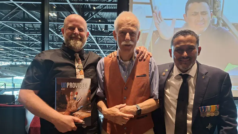 Drie mannen staan samen, een houdt het boek "UNPROVIDED" vast, achtergrond met vliegtuigen en een groot scherm. 