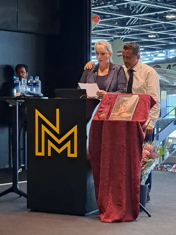Een vrouw en man staan achter een podium met een rode doek, microfoon en bloemen. Achtergrond: vliegtuig en NMM-logo.