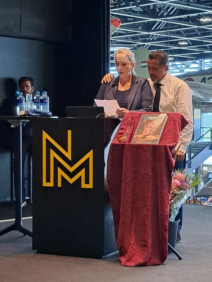 Een vrouw en man staan achter een podium met een rode doek, microfoon en bloemen. Achtergrond: vliegtuig en NMM-logo.