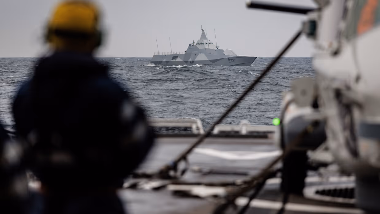 Een militair schip is zichtbaar op de horizon, op de voorgrond staat een schipmedewerker.