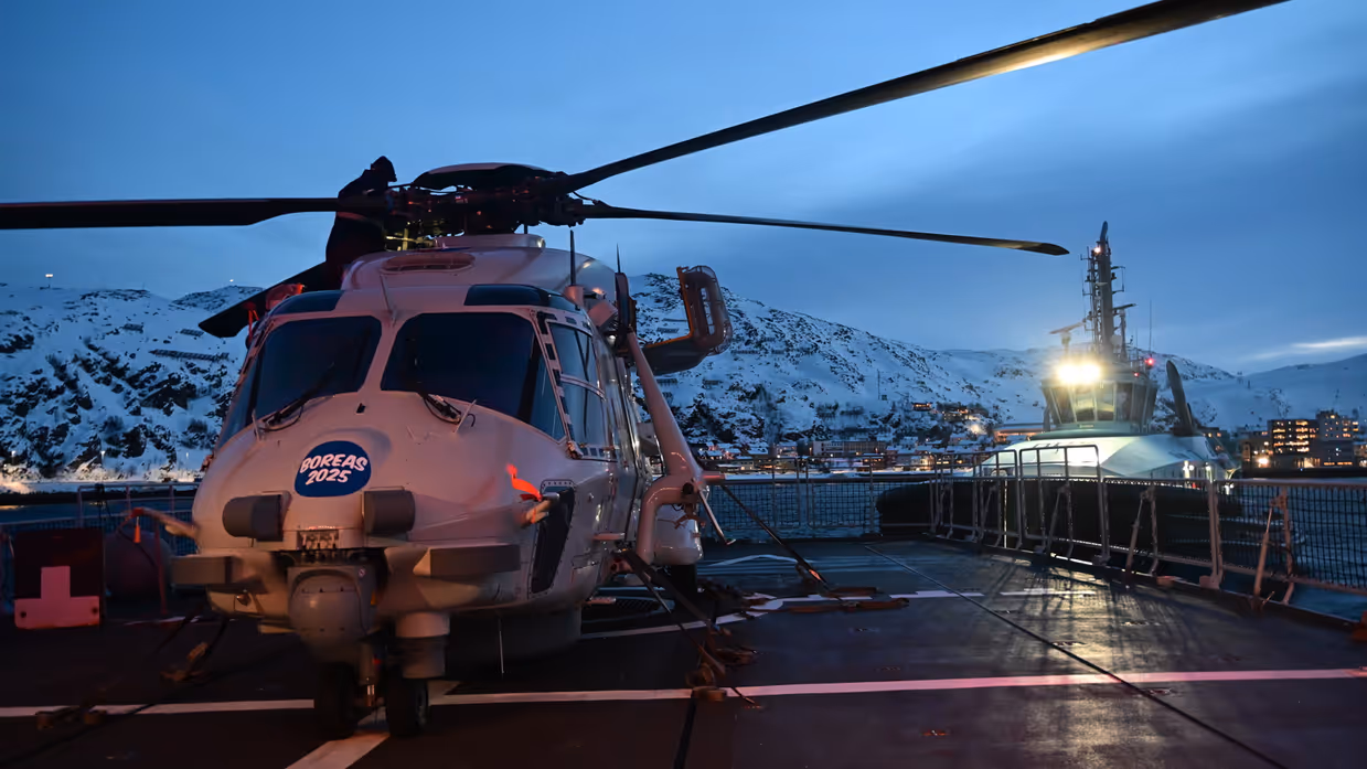 Helikopter op een landingsbaan met een militair schip in de achtergrond.