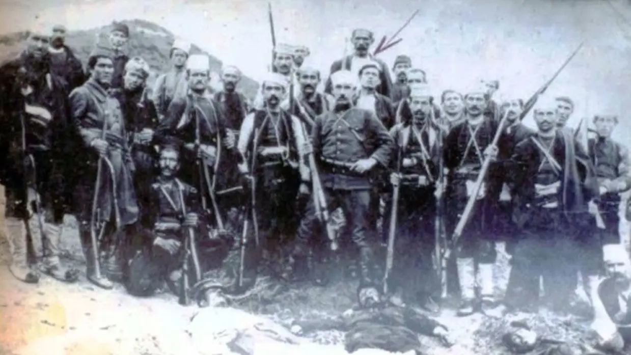 Een oude zwart-witfoto van een groep gewapende mannen in traditionele kleding, poserend achter een liggend persoon.