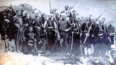 Een oude zwart-witfoto van een groep gewapende mannen in traditionele kleding, poserend achter een liggend persoon.