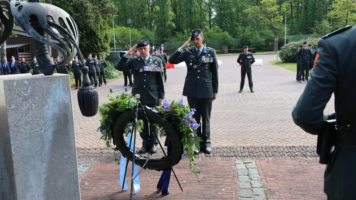 Twee militairen in uniform salueren bij een krans met bloemen en linten, omringd door andere militairen in een groene, open omgeving. 