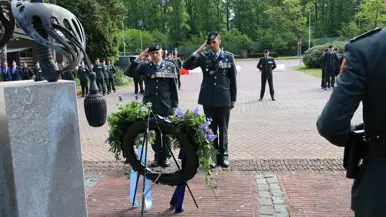 Twee militairen in uniform salueren bij een krans met bloemen en linten, omringd door andere militairen in een groene, open omgeving. 