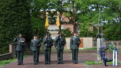 Vijf militairen in groene uniformen salueren in een rij, met vlaggen en een krans met bloemen op een stenen plein.