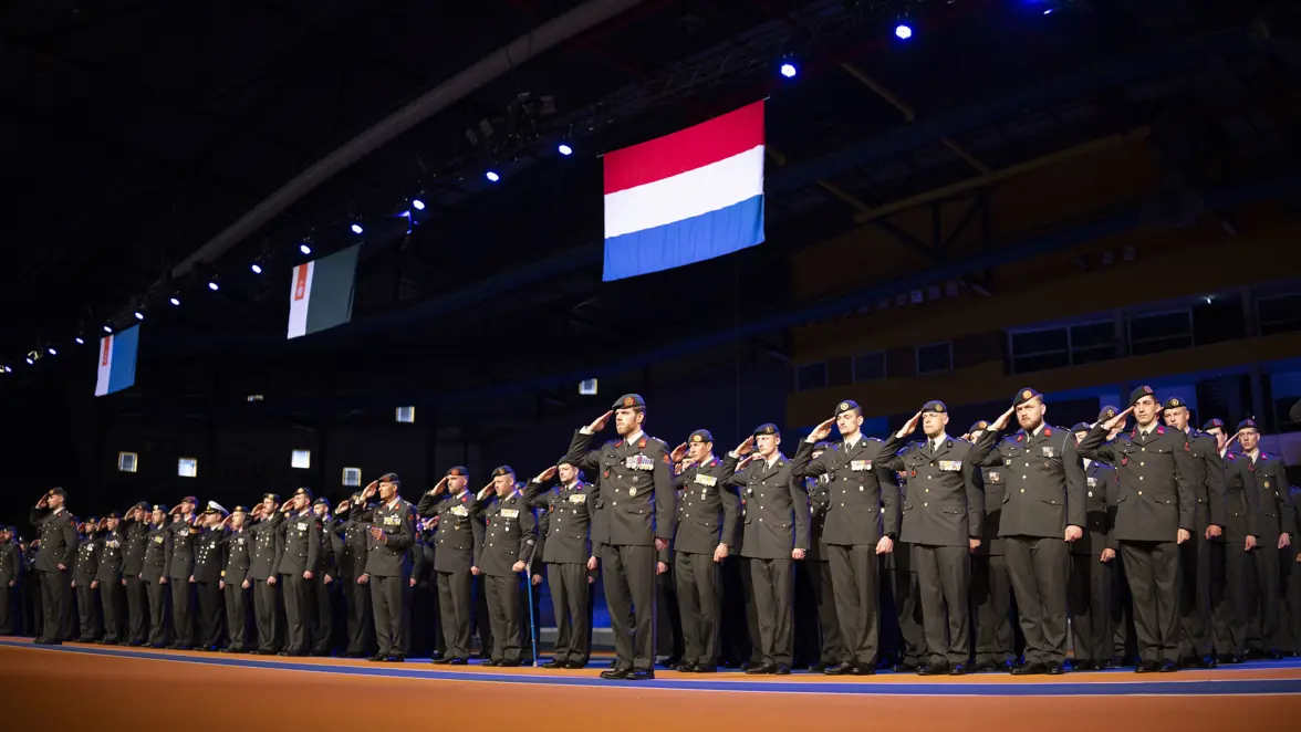 Militairen staan naast elkaar opgesteld terwijl ze salueren met boven hen de Nederlandse vlag.