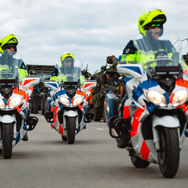 Motoren marechaussee rijden naast militaire voertuigen.