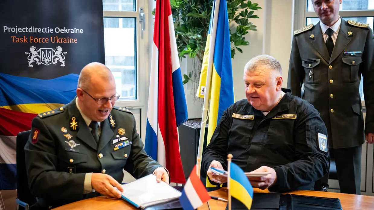 Kolonel Simon Wouda en chief engineer Vadym Nurmahomedov tekenen zittend aan een tafel de overdracht. Achter hen de Nederlandse en Oekraïense vlag.