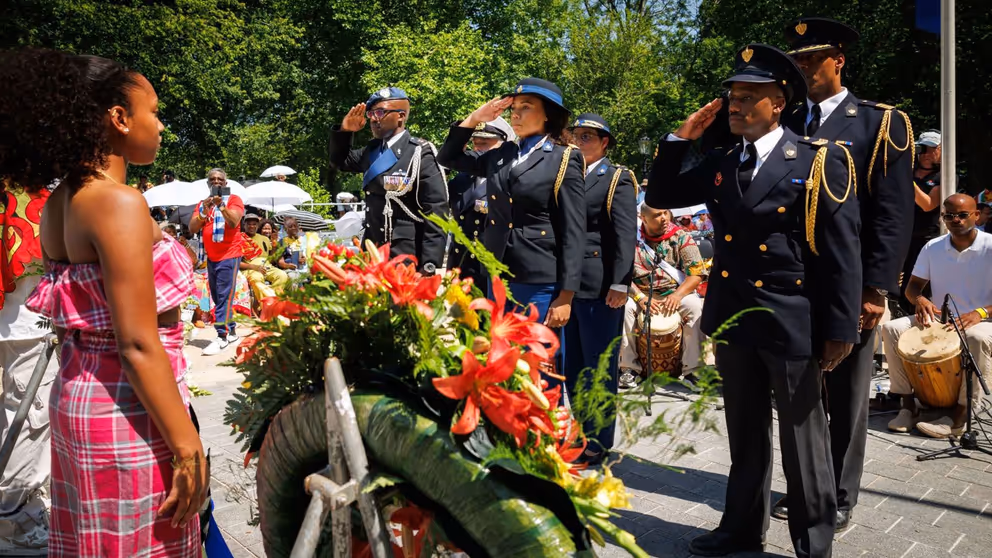 Militairen van het Multicultureel Netwerk Defensie salueren bij het Nationaal Monument Slavernijverleden. Een krans met bloemen op de voorgrond.