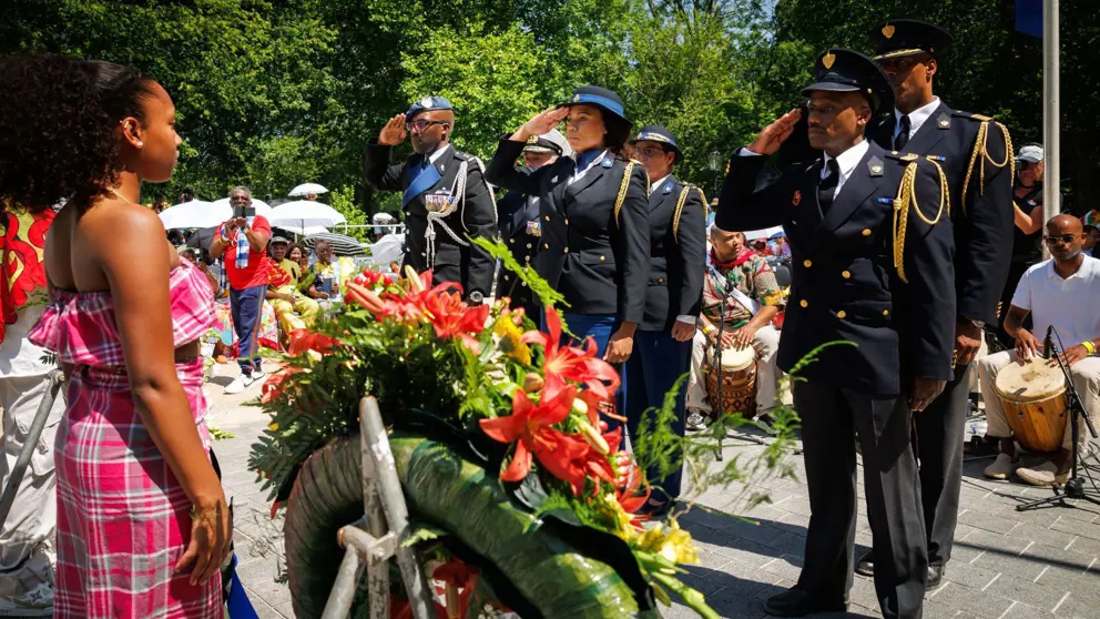 Militairen van het Multicultureel Netwerk Defensie salueren bij het Nationaal Monument Slavernijverleden. Een krans met bloemen op de voorgrond.