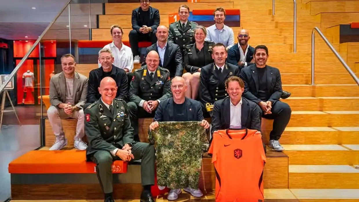 Groep KNVB'ers en mensen van Defensie op een tribune met een Oranje-shirt en een camouflage-shirt