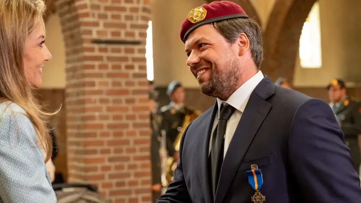 Een man in pak met een rode baret op zijn hoofd schudt lachend de hand van de minister.