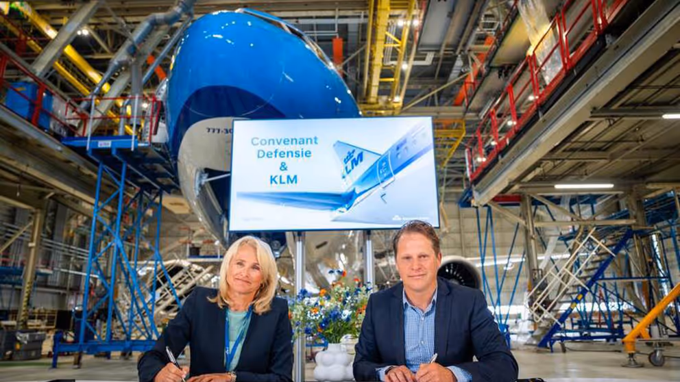 Vrouw en man achter tafel in vliegtuighangar ondertekenen samenwerkingsovereenkomst.