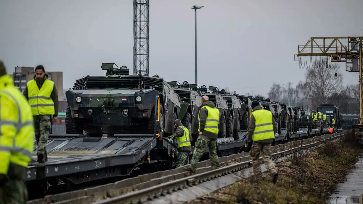 Legervoertuigen komen per spoor aan. Militairen met gele hesjes staan langs het spoor.