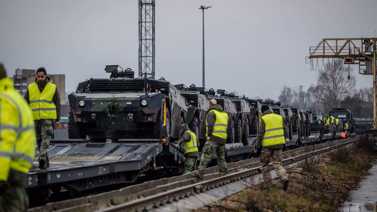 Legervoertuigen komen per spoor aan. Militairen met gele hesjes staan langs het spoor.