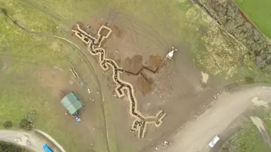 Links: Luchtfoto van loopgraven op militair oefenterrein. Rechts: Militair met wapen hurkt in loopgraaf waarvan de wanden met golfplaten zijn bekleed. Op zijn helm een bos stro als camouflage.