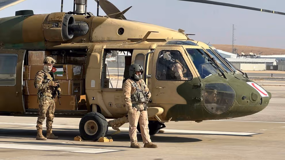 Dozen met hulpgoederen staan in een helikopter met de deur open. 2 militairen staan naast de helikopter..