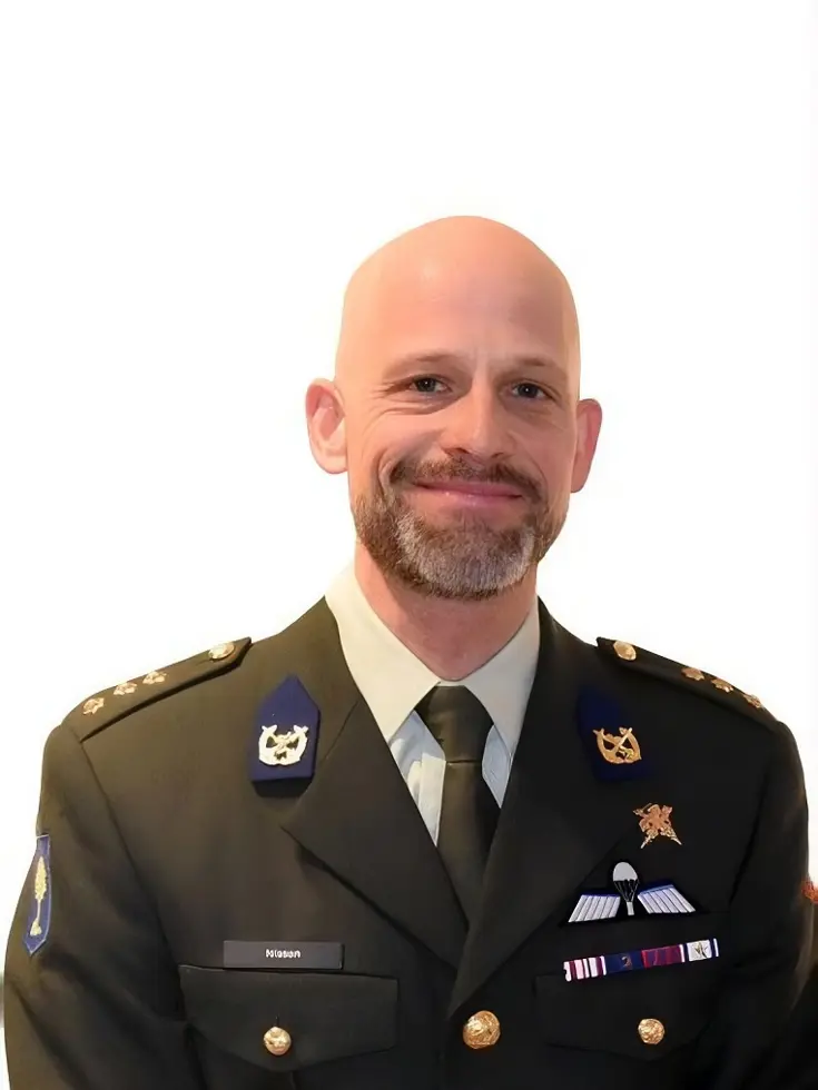 Een glimlachende, kaalgeschoren man in militair uniform met onderscheidingen, insignes, en een das, draagt op zijn borst een naamkaartje met ‘Nissen’. 