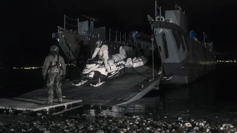 Een marinier in wit pak bestuurt een sneeuwscooter. Hiermee verlaat hij, in het donker, een landingsvaartuig.
