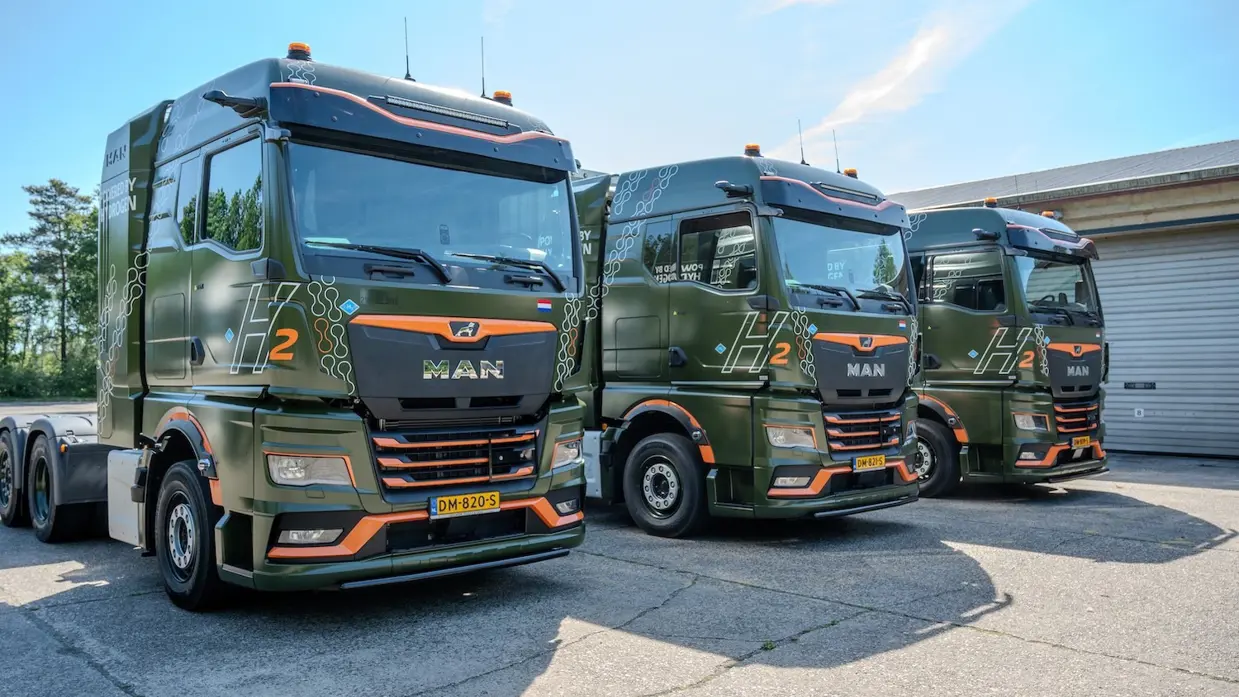 Drie groene trucks op een rij, met oranje accenten. Op de cabine staan de lettercijfercombinatie H2, voor waterstof.