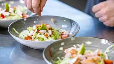 Op de linkerfoto voegen twee handen ingrediënten toe aan salades. Op de rechterfoto kijkt generaal Jobse toe hoe één van de cursisten een stuk groente snijdt. 