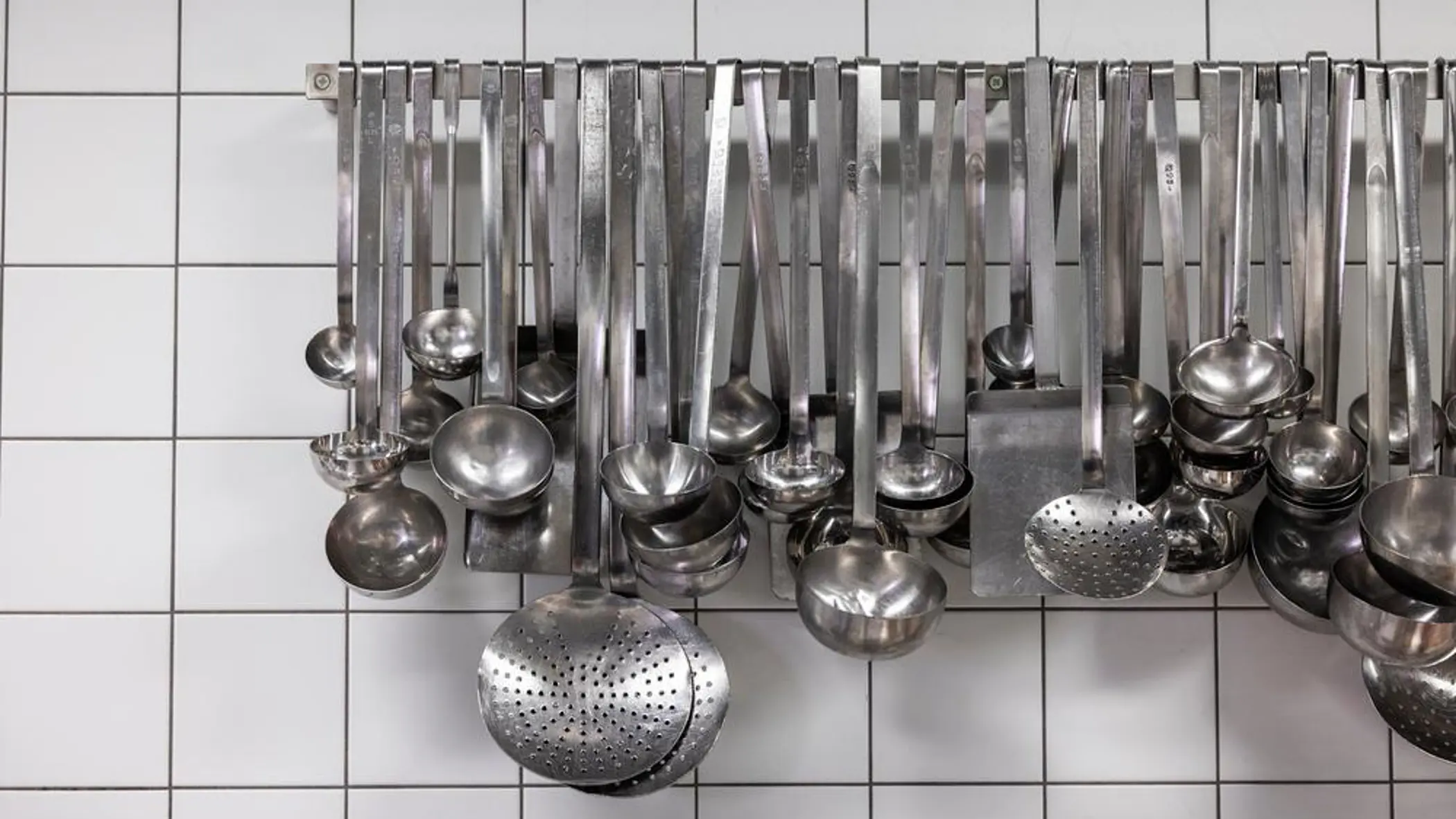 Grote lepels en ander staalkleurig keukengerei hangt aan een rek op een witte tegelmuur. 