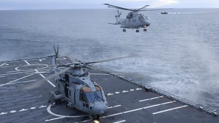 Een helikopter vliegt boven landingsdek schip. Een ander toestel staat op het dek.